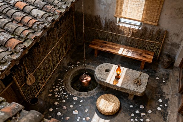 Overhead view of a small wabi-sabi courtyard patio with weathered textures: a moss-flecked terracotta-tile roof, reed-and-bamboo fencing, and rough plaster walls. A rough stone table holds an amber oil lamp beside dried stems; a circular stone well or fire pit suspends a copper kettle on a chain, a low solid-wood bench sits under a rolled bamboo shade, seashells scatter across worn concrete, and a round woven straw cushion rests in a sunbeam.