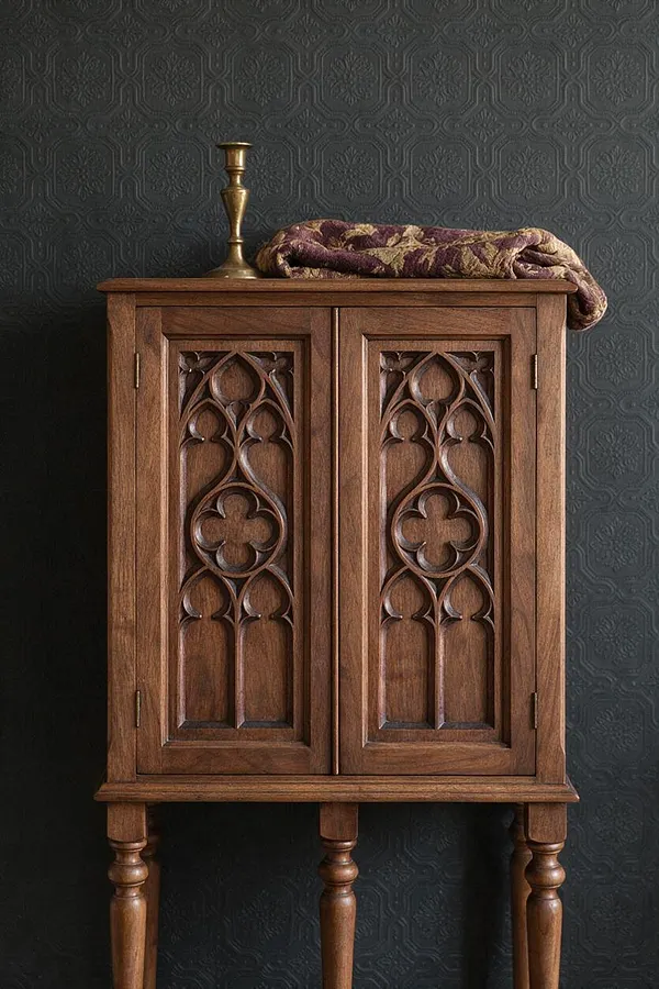 A small Gothic-style wooden cabinet stands against a dark embossed wallpaper backdrop. The cabinet has two carved doors with pointed tracery arches, turned legs, and a warm brown finish, with a brass candlestick and folded patterned textile placed on top.