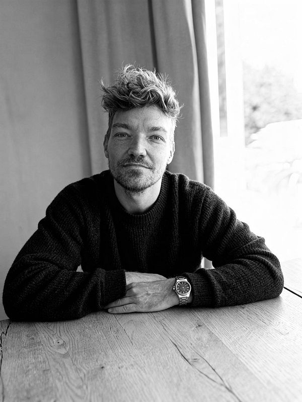 A man with tousled hair sits at a wooden table, wearing a black sweater and a watch, in a black and white photo.