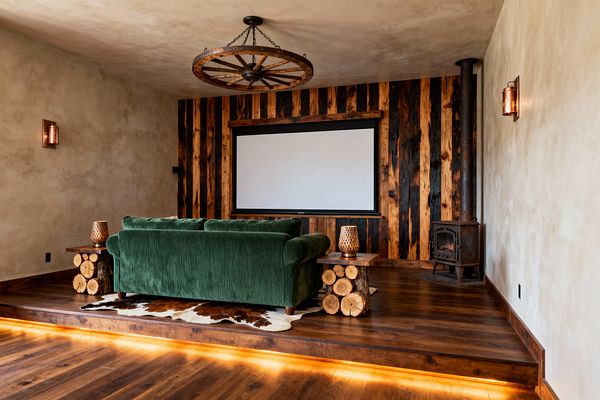 A rustic media room with a large projection screen set in a vertical accent wall of alternating charred and honey-toned wood planks. A forest-green velvet sofa sits on a brown-and-white cowhide rug, flanked by log-base side tables with perforated copper lamps. The walnut platform floor is underlit with warm LEDs. A wagon-wheel ceiling fixture and a black cast-iron wood stove sit against beige plaster walls, creating a cozy, cabin-like feel.
