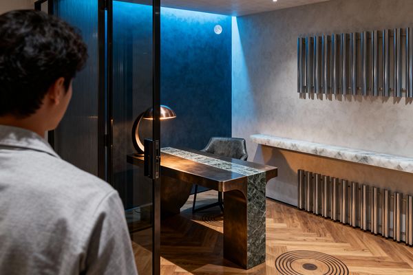 A person stands outside a glass-walled home office looking in at a sculptural bronze desk with a green marble stripe and a marble waterfall leg, lit by a dome lamp with a circular base. The room has a deep blue accent wall washed with cove LEDs and a small moon motif, pale gray plaster on adjacent walls, a floating white marble console above ribbed metal details, and warm herringbone wood flooring with a circular inlay.