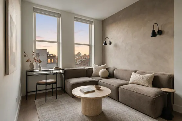 A modern studio apartment living area with a taupe sectional sofa against a textured beige wall, paired with black wall sconces and a slim black console desk by two tall windows. A round pedestal coffee table, woven rug, and soft neutral pillows create a calm, minimal atmosphere with sunset light and city views.