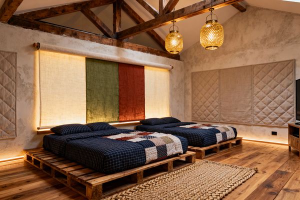 A farmhouse-style media room with two low beds set on reclaimed wood pallets over wide-plank floors. Exposed timber trusses, woven rattan pendants, and limewashed plaster walls frame navy grid bedding with patchwork quilts, a chunky jute rug, and linen wall panels, including a backlit roll-down in cream, olive, and rust. Quilted beige panels and a wood media console with TV complete the cozy, softly lit space.