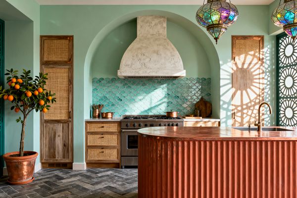 A Bohemian kitchen with mint-green walls features an arched alcove housing a plaster range hood and stainless stove, backed by glossy turquoise fish-scale tile. Natural wood cabinets with woven rattan fronts flank the range, while a fluted terracotta-toned island with a copper top and brass faucet sits on a charcoal herringbone floor. Jewel-toned Moroccan pendant lights and a teal lattice screen cast patterned sunlight; a potted orange tree adds warmth.