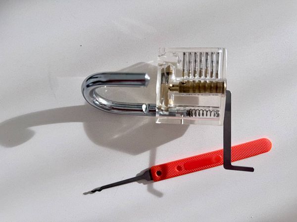 A transparent padlock with visible internal mechanism next to a red-handled lockpick tool.