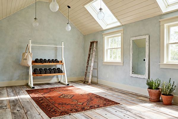 Bright farmhouse home gym with wide reclaimed wood floors, pale blue limewashed walls, and a cream shiplap ceiling with two skylights. A white metal rack with wood shelves holds black dumbbells, with a canvas tote hanging on the side. A rustic ladder leans near two double-hung windows, and a large distressed white mirror hangs on the wall. An orange-red patterned rug centers the room, with terracotta potted plants in the corner and glass pendant lights overhead.