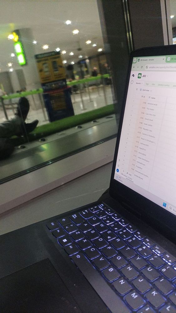 A person working on a laptop and taking notes at an airport terminal lounge with large windows showing an overcast sky and runway outside.