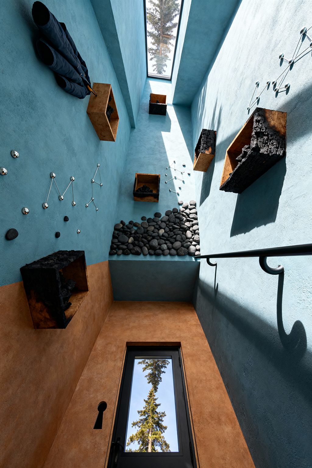 A tall mudroom with textured teal and ochre plaster walls features scattered wooden boxes holding charred timber, chrome ball-and-rod hooks arranged like constellations, and a ledge piled with smooth black river stones. Light from a ceiling skylight and a black-framed glass door casts long shadows across a black metal handrail and a pair of suspended black boots, creating a surreal, gravity-defying scene.