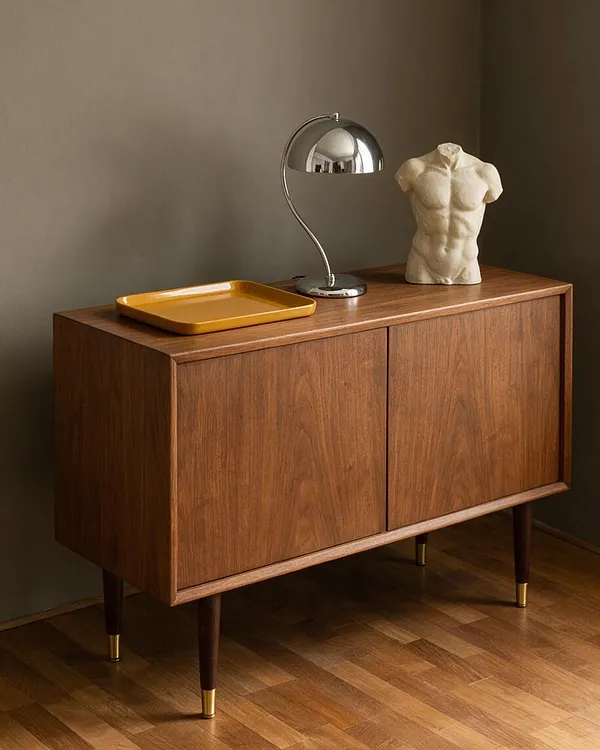 A walnut sideboard with clean mid-century lines stands against a muted taupe wall, topped with a chrome desk lamp, a mustard tray, and a small classical torso sculpture. The piece sits on tapered legs with brass caps over a warm herringbone wood floor.