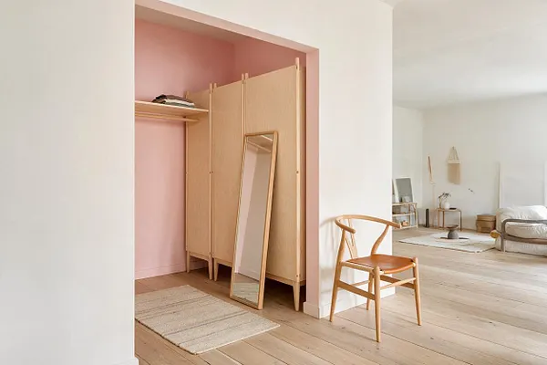 A Scandinavian-style closet nook with pale oak cabinet doors, a slim leaning mirror, and a blush pink back wall. The space opens into a bright minimalist room with light wood floors, a woven rug, and a wooden chair in the foreground.