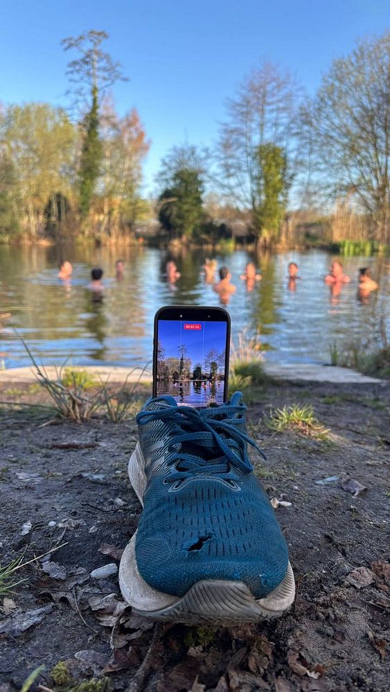 A serene waterscape shows several people swimming and floating in calm lake or sea waters near buoys and surrounded by forested hills under a cloudy sky.