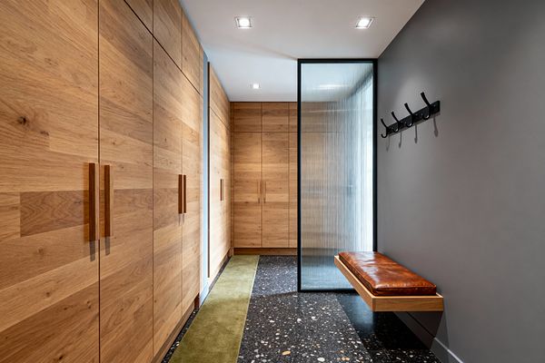 A contemporary mudroom with full-height oak cabinets on the left and at the far end, each with long vertical wood pulls. On the right, a charcoal-gray wall holds a row of black hooks above a floating oak bench with a cognac leather cushion. A black-framed, fluted-glass partition screens light. The floor is dark terrazzo with pale speckles and a narrow olive runner. Recessed ceiling lights illuminate the clean, streamlined space.
