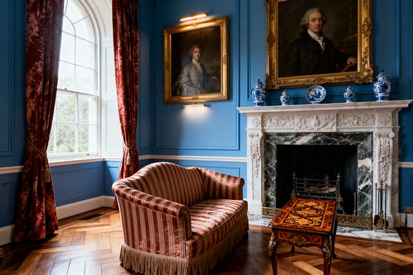 A traditional living room with saturated blue paneled walls features an ornate white carved marble mantel around a dark green marble fireplace. Blue-and-white porcelain sits on the mantel beneath two gilded portrait paintings lit by brass picture lights. A striped rose-red settee with bullion fringe faces an inlaid marquetry table on a herringbone wood floor. An arched window with red velvet drapes and tassel tiebacks admits daylight; brass fireplace tools rest by the hearth.
