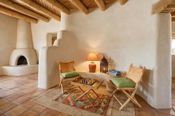 A Southwestern-style game room with white adobe walls, rounded built-in fireplace, exposed log ceiling beams, and terracotta tile flooring. Two bamboo-framed chairs with green cushions face a small wooden chess table on layered woven rugs, with a warm table lamp and decorative lantern creating a cozy atmosphere.