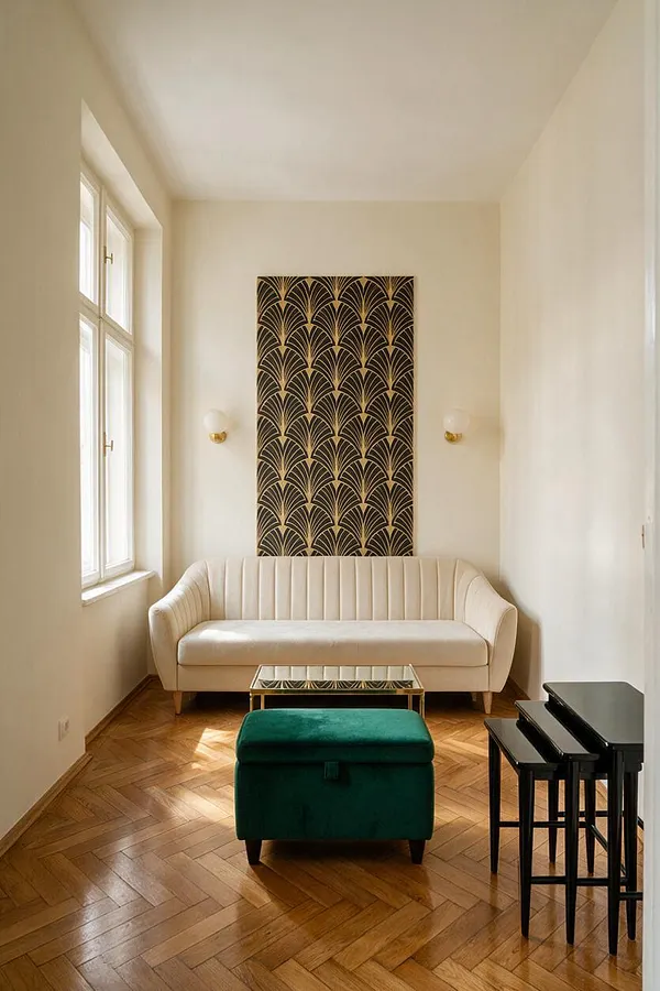 A small studio apartment living area with cream walls, a white upholstered sofa, and a dark Art Deco wall panel centered above it. A green velvet ottoman sits in front of a mirrored brass coffee table, while nested black side tables stand to the right. Natural light enters through tall windows on the left, highlighting the herringbone wood floor.