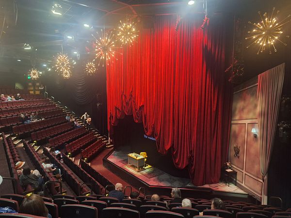 The image captures the interior of a theater with a red curtain and audience seating.