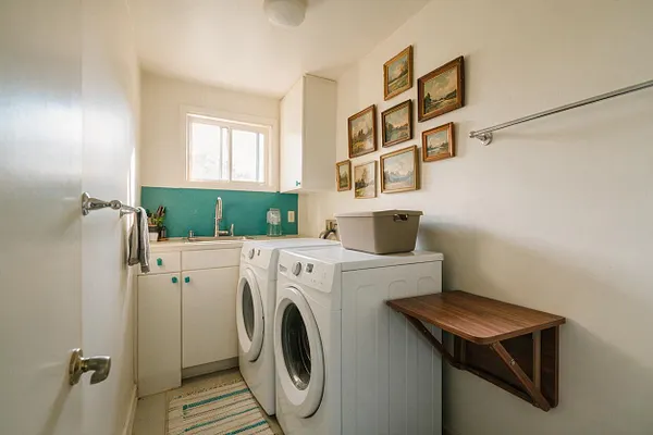 A compact laundry room with a white washer and dryer side by side under a small window. The room features a teal backsplash behind a sink, cream cabinets, framed artwork arranged on the wall, a wall-mounted folding shelf, and a striped rug on the floor.