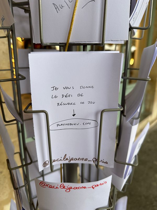 A collection of white cards displaying a challenge related to a game, attached to a metal stand in Paris.