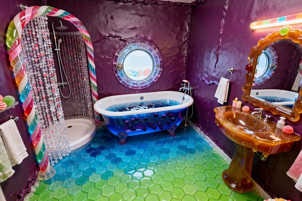 A playful bathroom features glossy purple walls, a freestanding translucent blue tub with purple orb accents below a round porthole window, and a shower framed by a striped candy-cane arch. The shower uses a curtain of clear and colored beads. The floor is a gradient of shiny hexagon tiles shifting from lime green to aqua and cobalt. At right, an amber, caramel-like resin pedestal sink with a scalloped mirror is lit by a multicolor vanity light, with chrome fixtures and white towels.