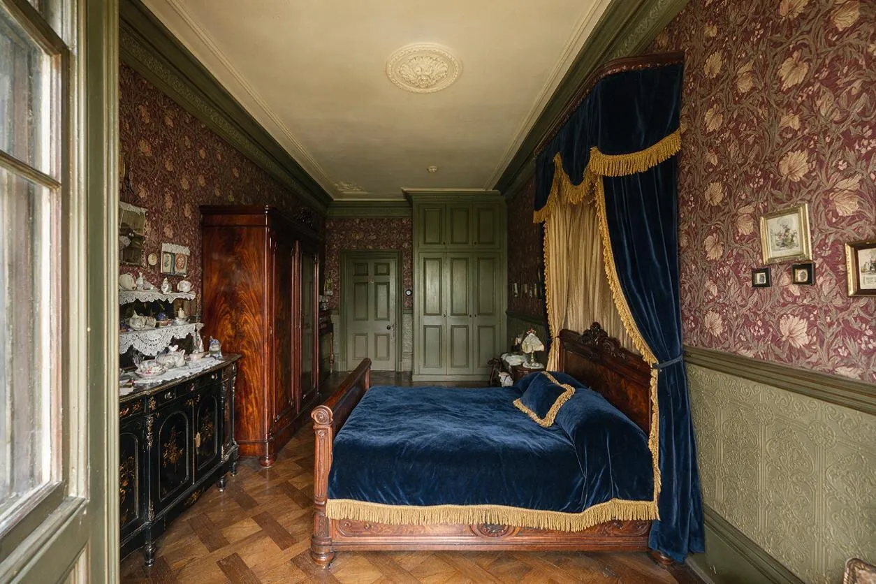 A narrow Victorian-style guest bedroom with a carved wooden bed dressed in deep blue velvet and gold fringe, framed by heavy blue and gold drapery. The room features patterned floral wallpaper, olive green trim, parquet wood flooring, a black painted cabinet with decorative dishes, and a tall wooden wardrobe.