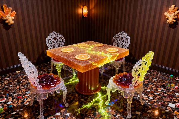 Playful dining vignette featuring a square orange gummy-like table dusted with sugar texture and citrus slice inlays, glowing from within over a ribbed pedestal with neon-yellow cracks that spill onto the floor. Four translucent chairs with rock-candy style backs and glossy jelly seats sit on a multicolor terrazzo floor, against chocolate-brown striped walls lit by warm amber bubble sconces.