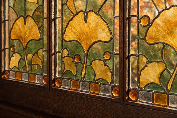 A close-up of a Craftsman-style stained glass window insert featuring repeating ginkgo leaf motifs in amber, gold, and green glass. Dark lead came outlines the organic pattern, and a dark stained wood frame anchors the bottom edge.