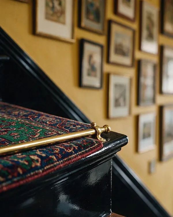 A close-up of a black-painted staircase with a richly patterned red, green, and blue stair runner secured by a polished brass rod. In the background, a warm mustard wall is covered with a gallery of framed artwork, creating a layered maximalist look.