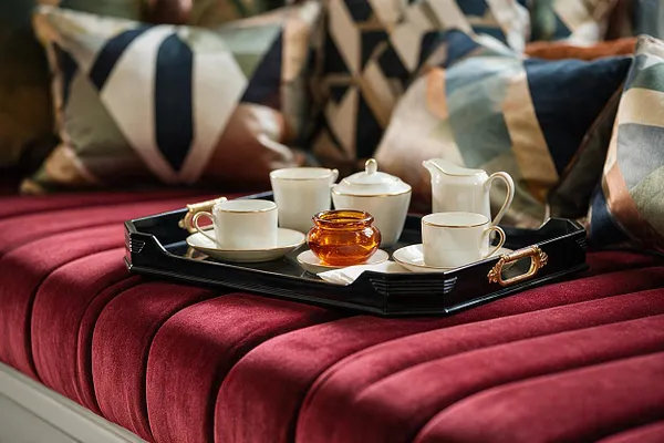 A plush burgundy velvet banquette is styled with a black lacquer tray holding a white porcelain tea set and amber glass candle or jar. Behind it, oversized cushions display bold geometric patterns in cream, navy, sage, blush, and copper, reinforcing the Art Deco-inspired look.