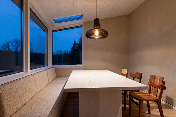 A Scandinavian dining nook with an L-shaped built-in bench upholstered in cream bouclé sits beside large corner windows and a roof skylight. A light pine table faces three walnut chairs with cognac leather back straps, set against warm beige plaster walls and a wood-clad ceiling. A smoked glass pendant with a visible bulb provides soft, warm light, contrasting with the cool blue evening view outside.