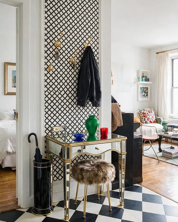A bright studio apartment entry nook with a mirrored console table framed in brass, set against a black lattice wall panel. Gold hooks hold a dark coat above a fur-topped stool, and a black umbrella stand sits beside the table on a bold black-and-white checkered floor. The living area beyond includes white walls, colorful pillows, and warm wood flooring.