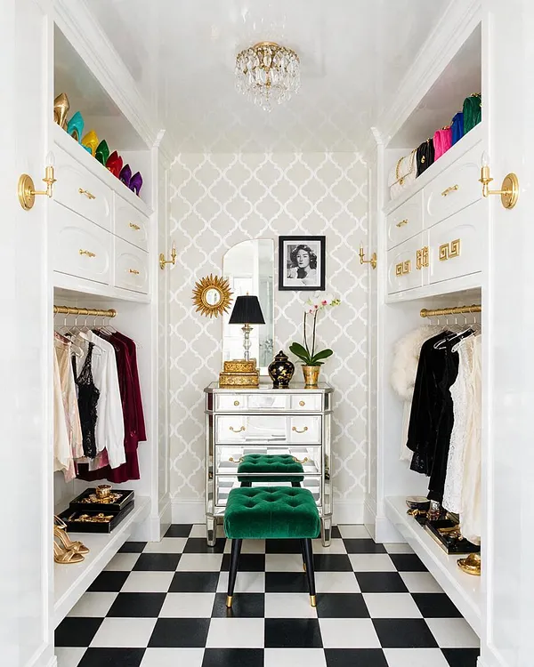 A narrow Hollywood Regency closet with white built-in shelving and drawers, gold hardware, and a black-and-white checkered floor. A mirrored chest, green velvet stool, crystal chandelier, and colorful accessories add a luxe, polished look.
