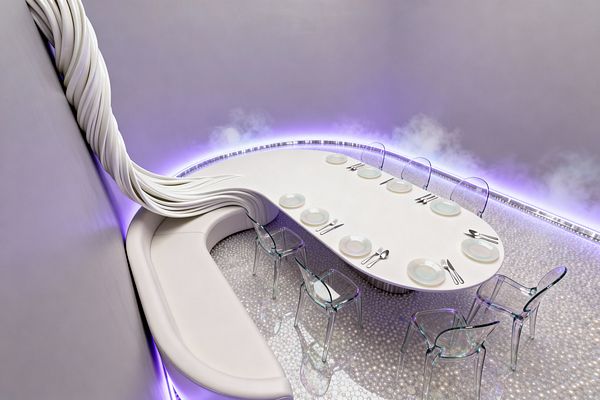 A futuristic dining room with a kidney-shaped white table integrated into a curved banquette. Transparent polycarbonate chairs face the table, which is set with simple plates and flatware. A twisting white ribbon-like sculpture descends the wall into the seat. The floor has a pearlescent, bead-like texture that reflects violet LED cove lighting around the perimeter, with a light mist effect along the wall, creating an ethereal, high-tech atmosphere.