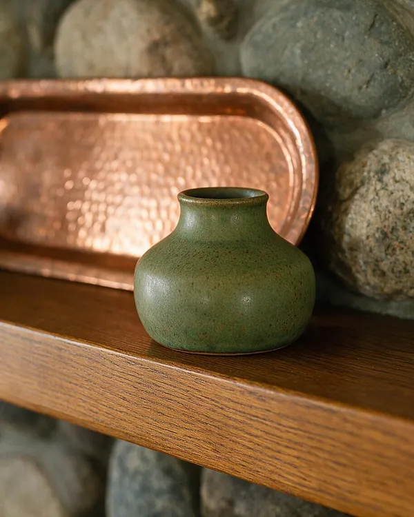A small, rounded green ceramic vase sits on a warm wood shelf in front of a hammered copper tray and a stacked stone backdrop. The composition highlights earthy textures and handcrafted materials typical of Craftsman-inspired decor.