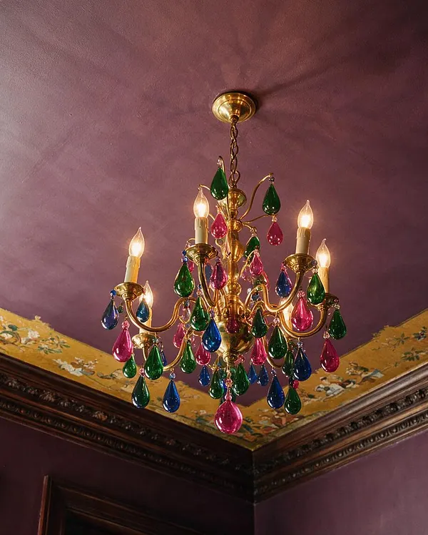 A gold-toned chandelier hangs from a plum purple ceiling in a richly decorated room. The fixture has candle-style bulbs and multicolored teardrop crystals in green, pink, and blue, with ornate trim and patterned wallpaper border visible along the ceiling edge.