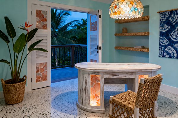 A tropical home office with seafoam walls opens to a balcony of palms through white louvered double doors. A curved, whitewashed wood desk features two backlit orange‑pink stone panels, while a dome pendant of multicolored discs glows above. A rope‑woven chair sits on speckled terrazzo flooring. Built‑in shelves wrapped in thick natural rope hold baskets and books, and an indigo tie‑dye textile hangs on the wall beside a potted bird‑of‑paradise.