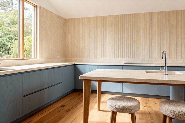 Light-filled Scandinavian kitchen with L-shaped base cabinets in muted blue-gray beneath pale beige stone countertops. A matching stone-topped, table-style island with an integrated sink and chrome gooseneck faucet stands on rounded wood legs beside two round bouclé stools. Vertically fluted beige wall cladding, honey oak plank flooring, a flush induction cooktop, and a wood-framed window to greenery complete the minimalist look.