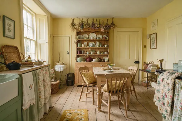 A cozy cottagecore kitchen with pale yellow walls, a wooden dining table and chairs, open hutch shelves filled with patterned dishes, and dried flowers displayed above. Lace, floral, and crocheted textiles soften the room, while natural wood floors, a large window, and vintage accessories create a warm country feel.