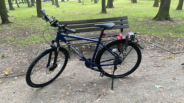 A new blue mountain bike parked near a bench in a park with trees and grass.