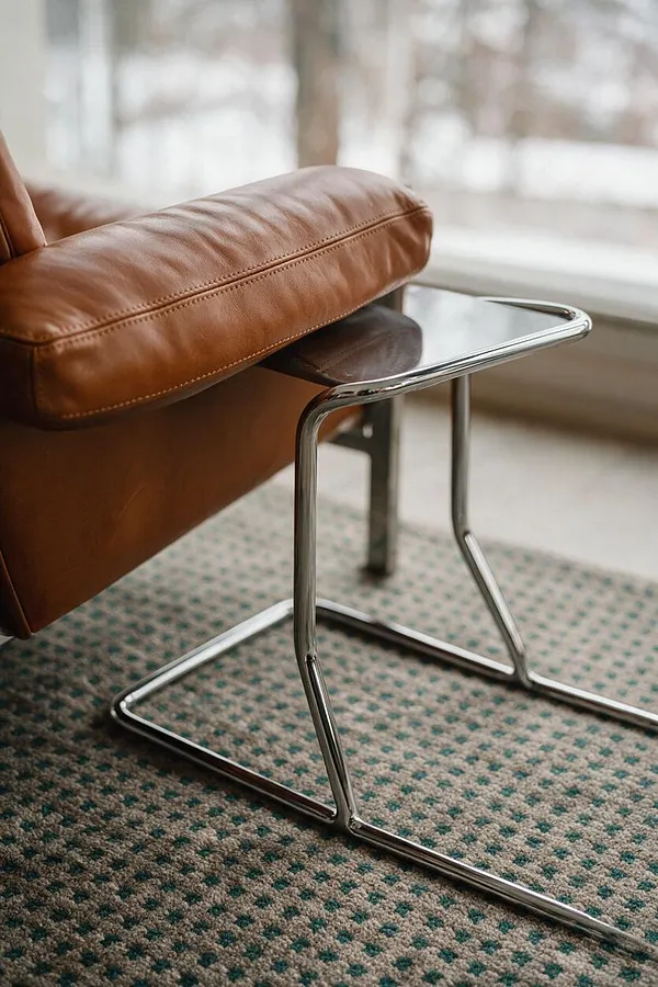 A close-up of a tan leather lounge chair showing a cushioned seat and polished chrome cantilever frame resting on a textured patterned rug. Soft daylight filters in from a window behind, creating a calm, modern eclectic look.