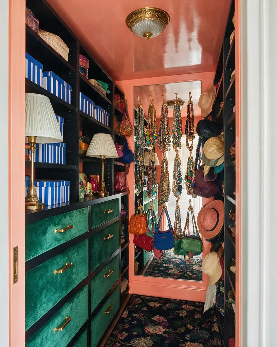 A narrow maximalist closet with coral-peach walls and glossy ceiling, black shelving, emerald velvet drawers, and gold hardware. Blue striped storage boxes, lamps, hanging hats, handbags, and layered jewelry fill the space around a large mirror and floral rug.