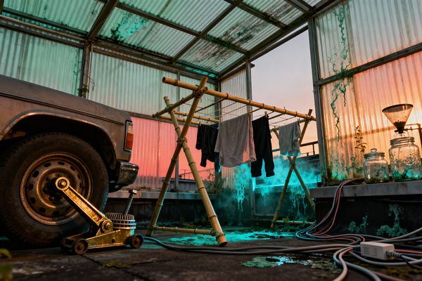 A makeshift laundry area sits inside a translucent corrugated shelter. A bamboo A‑frame rack strung with wire holds damp shirts and pants above a cracked concrete floor glowing with teal light and vapor; at left sits an old steel car and floor jack, while at right glass jars and a funnel apparatus rest on a ledge, with algae‑stained panels, coiled cables, and a pink‑orange sunset behind.