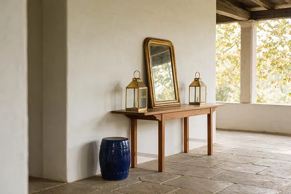 A covered exterior porch with white stucco walls and stone flooring features a simple wooden console table styled with a gold-framed mirror and two brass lanterns. A glossy navy ceramic stool sits beside the wall, while warm daylight and tree branches are visible through the open side.