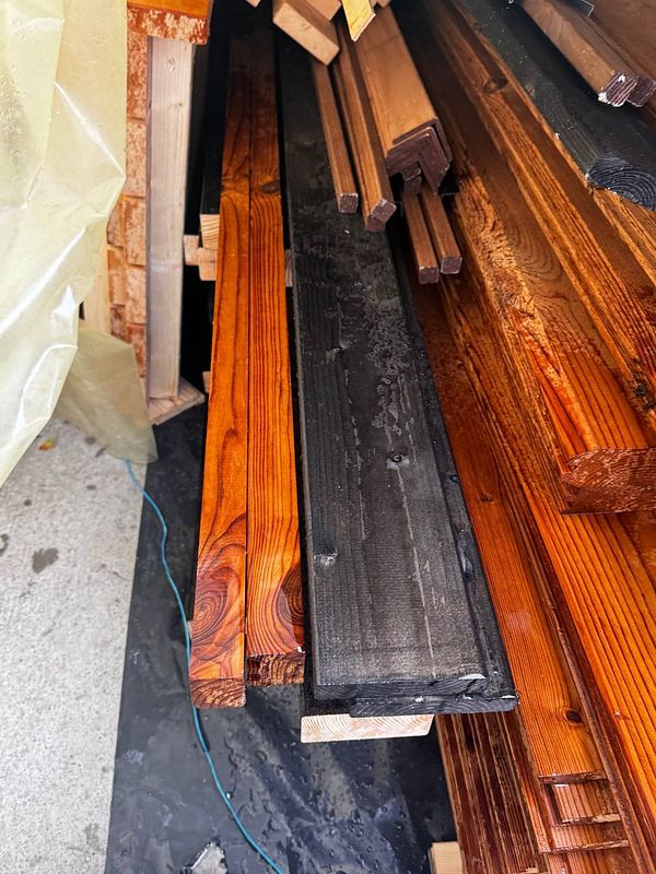 A collection of wooden planks, some wet, is stacked in a garden area.