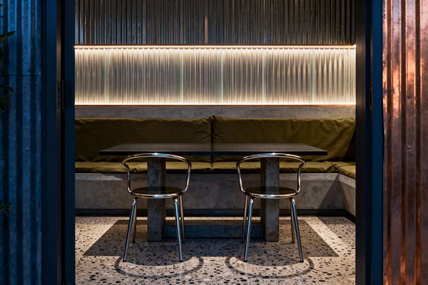 A compact industrial dining nook features an L-shaped concrete banquette with olive-green cushions facing a dark rectangular table on concrete plinths and two polished metal chairs. The back wall is clad in corrugated panels with a translucent ribbed band illuminated by warm LED strips, while a speckled terrazzo floor and an entry framed by blue-gray metal and copper cladding create a moody, intimate atmosphere.