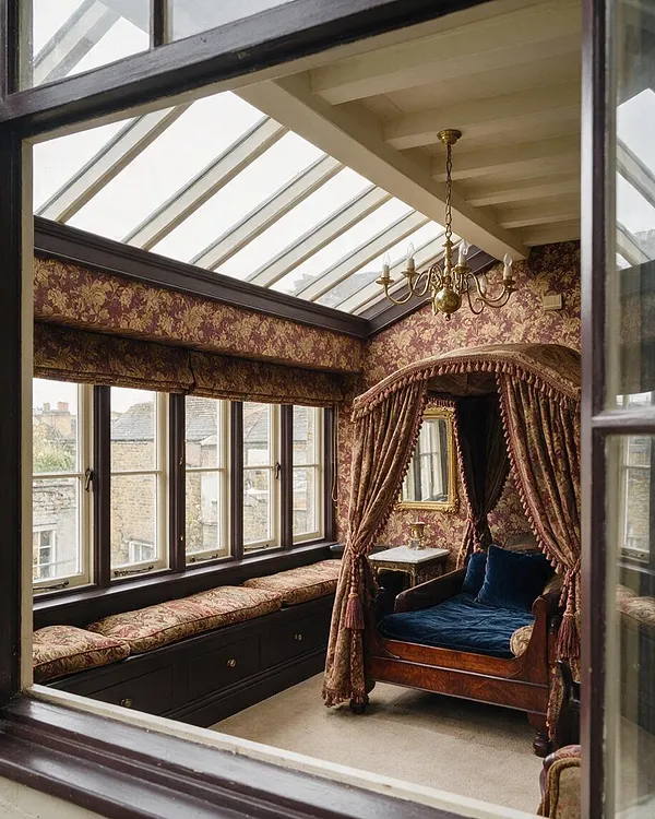 A Victorian-style sunroom with a glass roof, floral wallpaper, and dark wood window frames. The room features a built-in window seat with patterned cushions, a carved wooden daybed with a tasseled draped canopy, and a brass chandelier hanging from the ceiling.