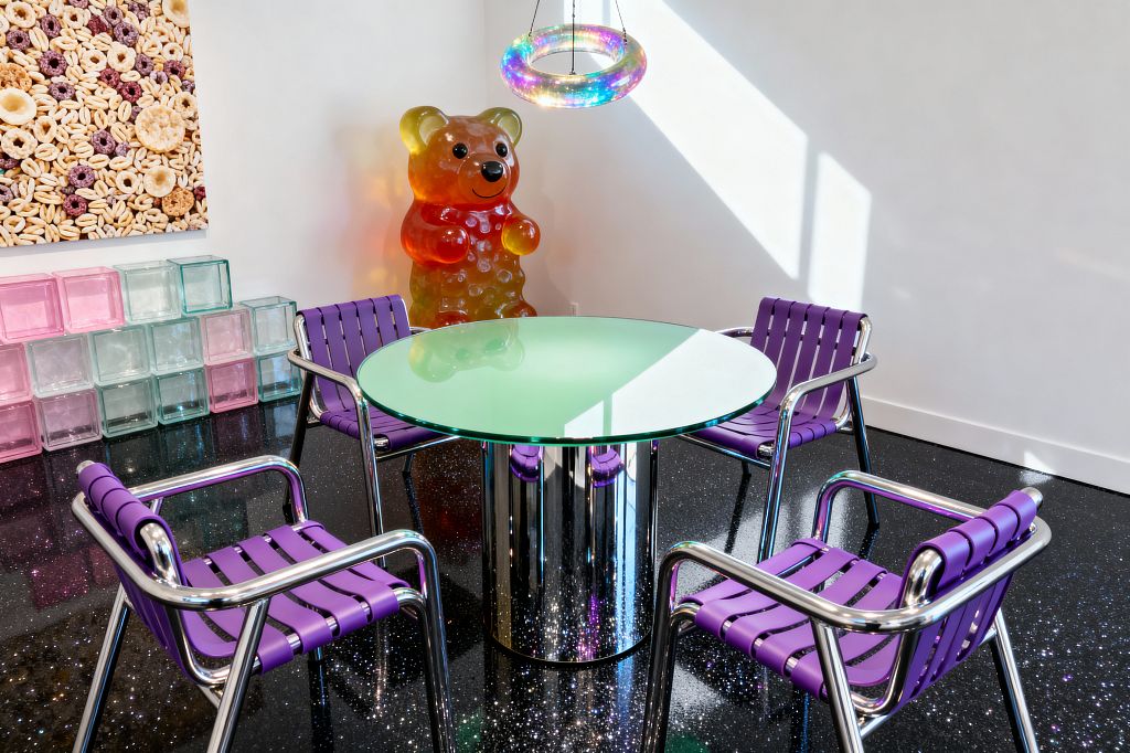 A Pop Art dining nook features a round, green-tinted glass table on a mirrored cylindrical base, surrounded by four chrome-framed chairs with purple strap seats; an iridescent ring pendant glows overhead, while a gummy bear sculpture and a large cereal photograph add playful scale. Stacked pink, mint, and clear glass blocks line the wall, with white walls and a glossy black, glitter-flecked floor illuminated by strong natural light.
