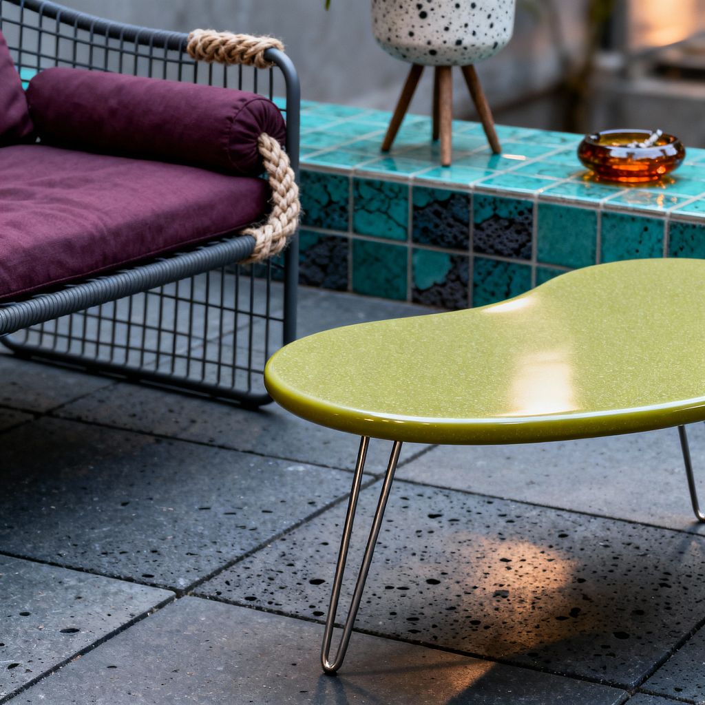 A mid-century patio vignette with an olive green, kidney-shaped coffee table on hairpin legs sitting on speckled charcoal concrete pavers. To the left is a lounge chair with a charcoal metal rod frame, deep plum seat and bolster cushions, and thick natural rope wrapped around the arm. Behind, a low ledge is faced in teal glazed tiles with dark variegation. On the ledge sit a speckled white ceramic planter on a small wooden tripod and a small amber glass dish.