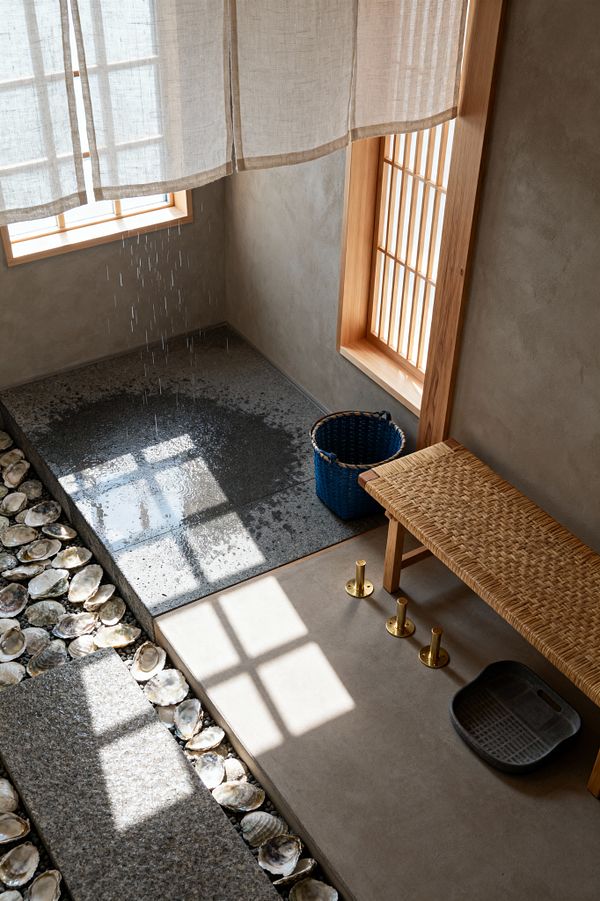 A serene Japanese-style mudroom features a raised plaster platform with a woven rattan bench, three floor-mounted brass pegs, and a dark rubber boot tray. Beside it, an indigo basket sits near a lower granite slab where water drips to rinse footwear, bordered by a channel filled with oyster shells and a textured stone tread. Light wood windows with slatted screens and a linen curtain filter soft natural light across the warm, minimal space.