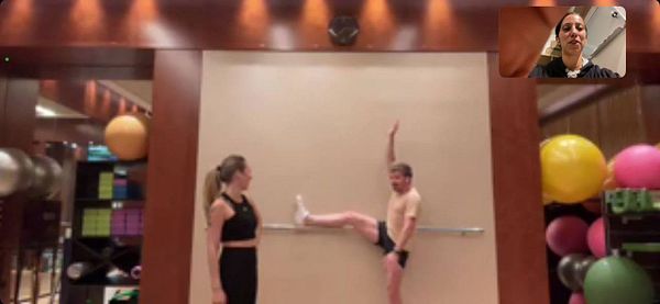 A virtual workout session is taking place in a gym setting with two individuals engaged in a stretching exercise.