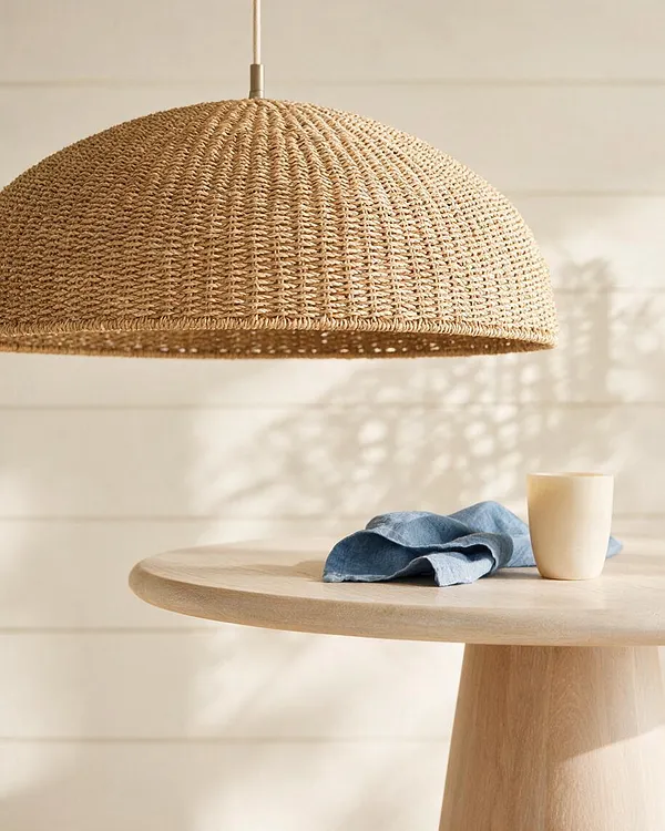 A coastal-inspired dining vignette featuring a large dome-shaped woven pendant light hanging above a pale wood round table. A soft blue linen napkin and a cream ceramic cup rest on the tabletop, while warm sunlight casts gentle shadows across a light paneled wall.